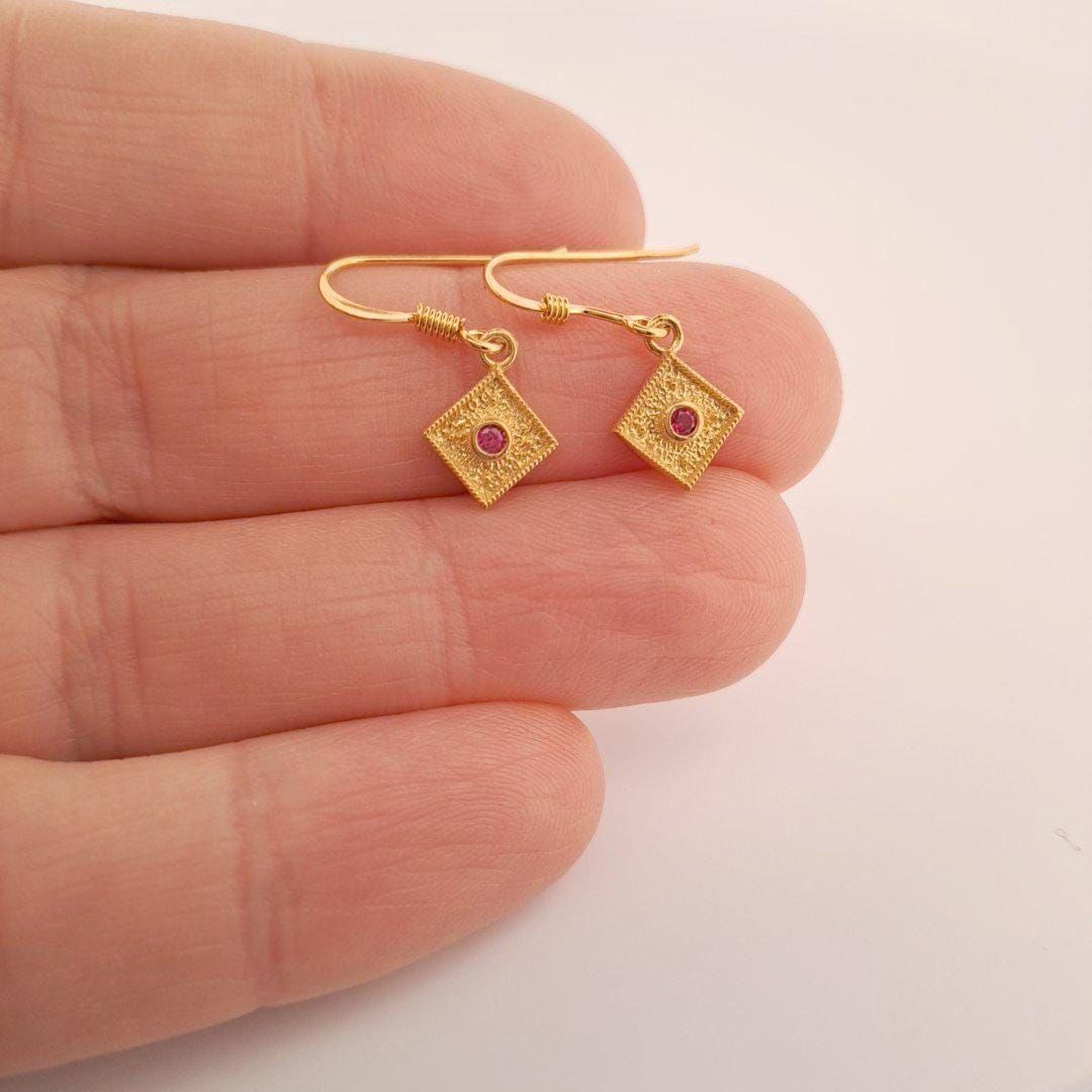 A pair of gold earrings with red gemstones is being held in a hand against a white background.