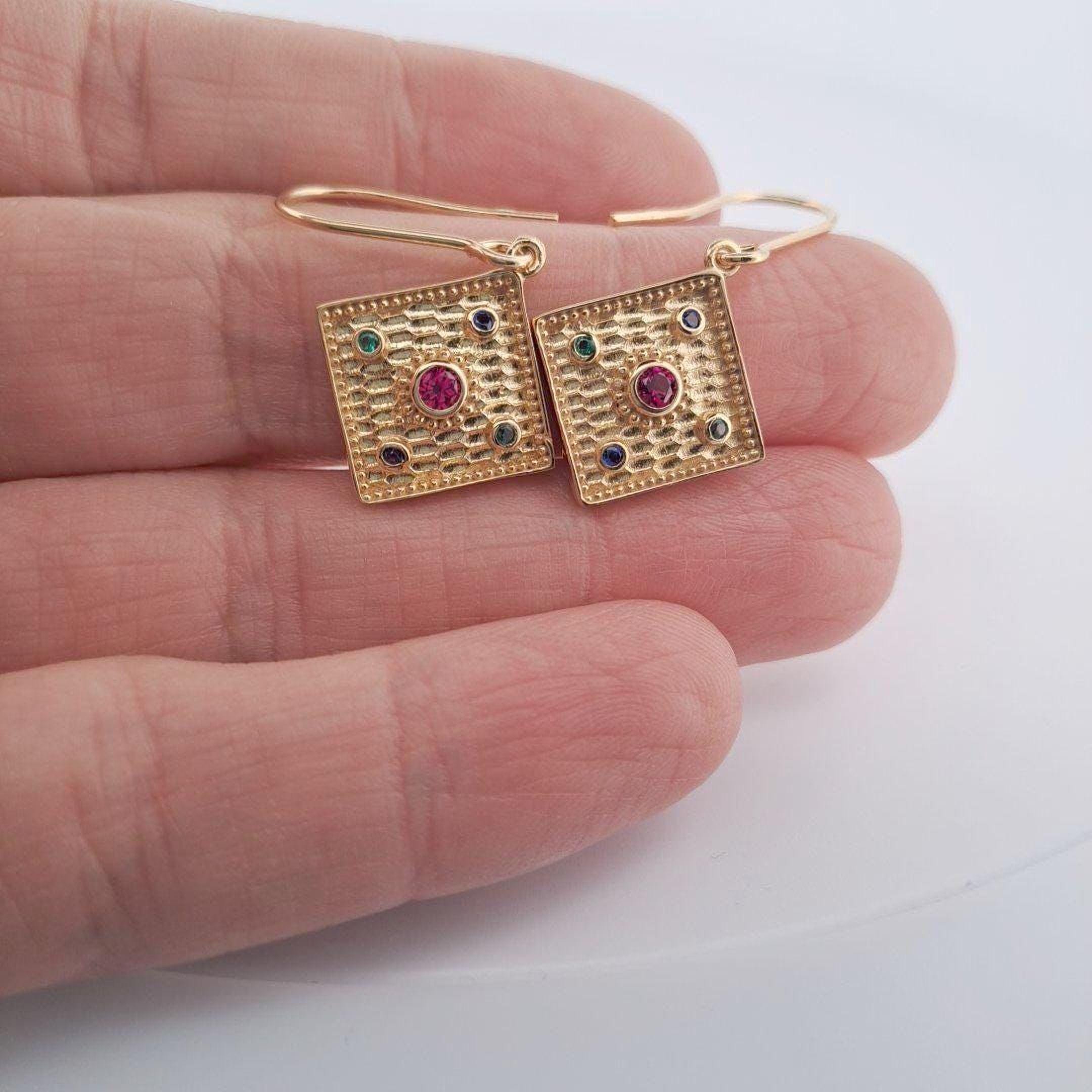 A pair of gold earrings with a square design and colorful gemstones, held in a person's hand.