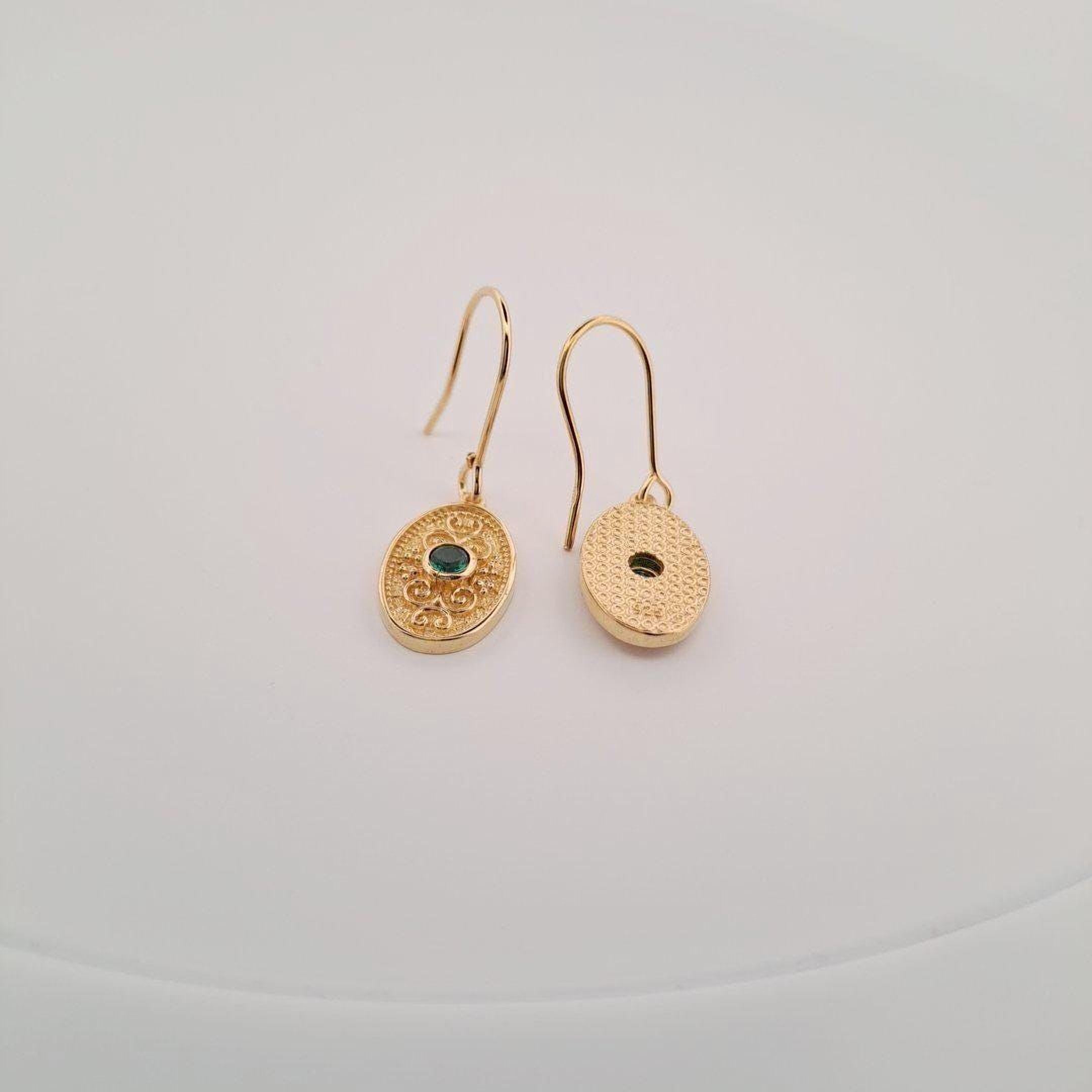 A pair of gold earrings with a green gemstone on one side and a round charm on the other, both hanging from hooks against a white background.