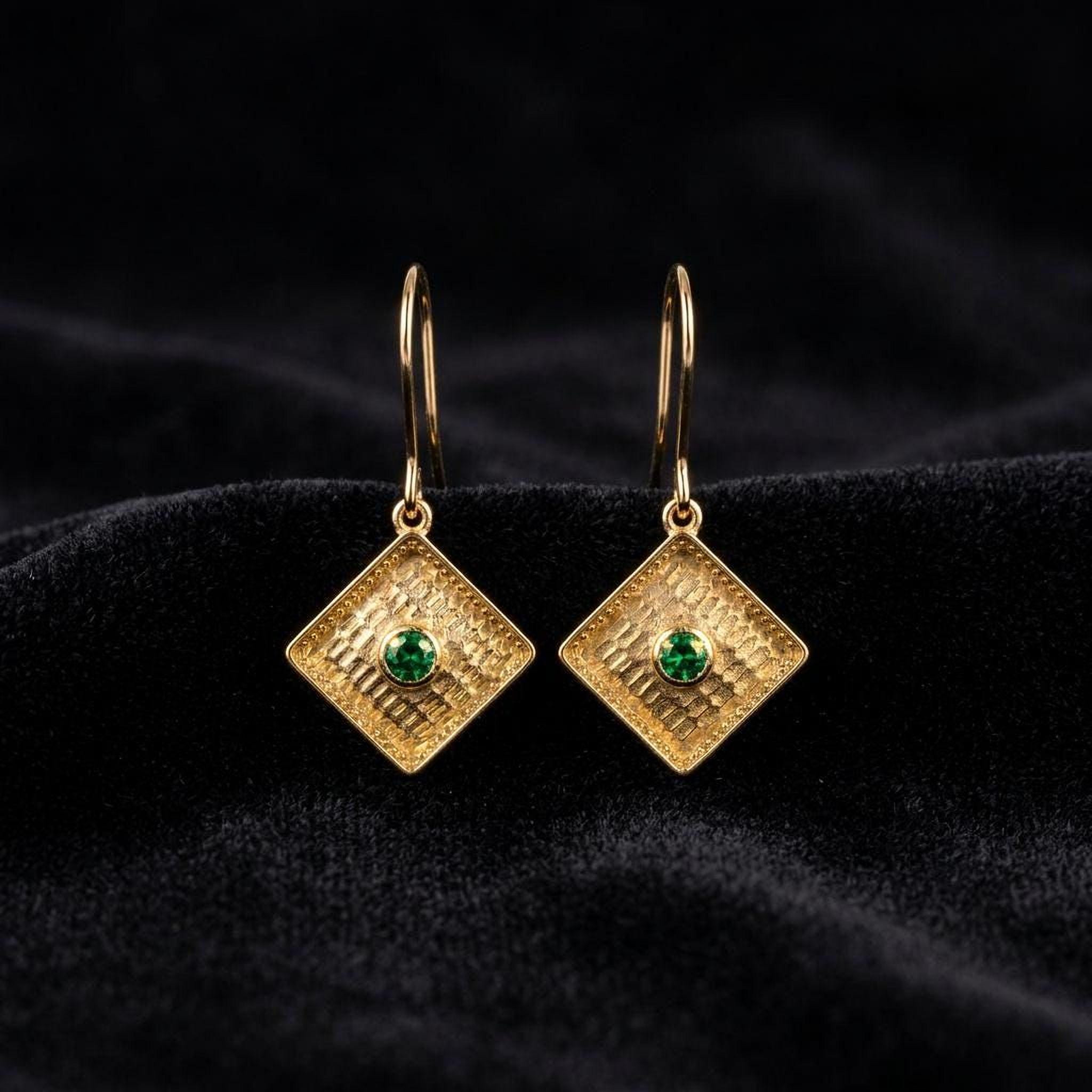 A pair of gold earrings with a green gemstone set in a diamond-shaped design, displayed on a black velvet background.