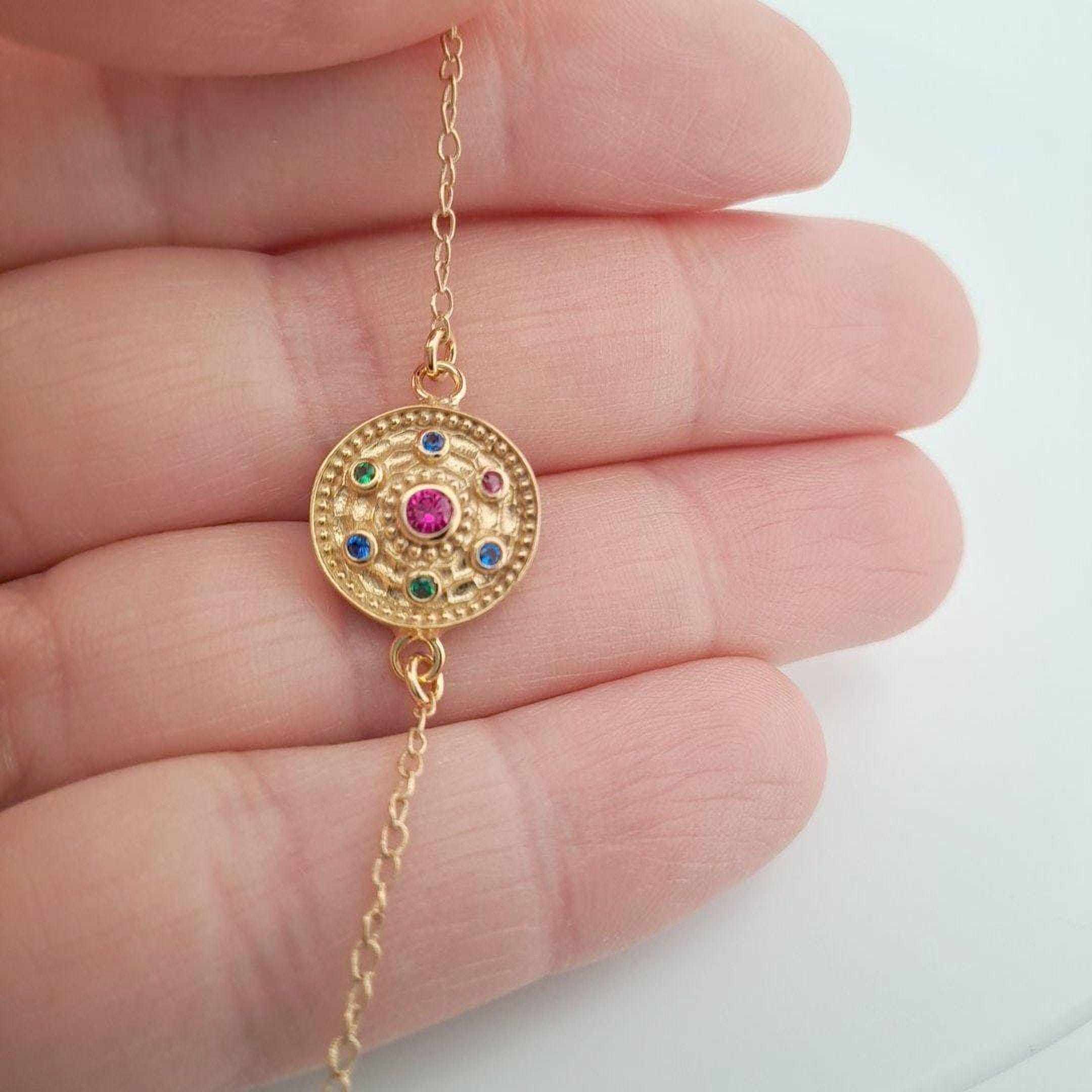 A hand holding a gold-colored necklace with a circular pendant featuring a red gemstone surrounded by smaller gemstones.