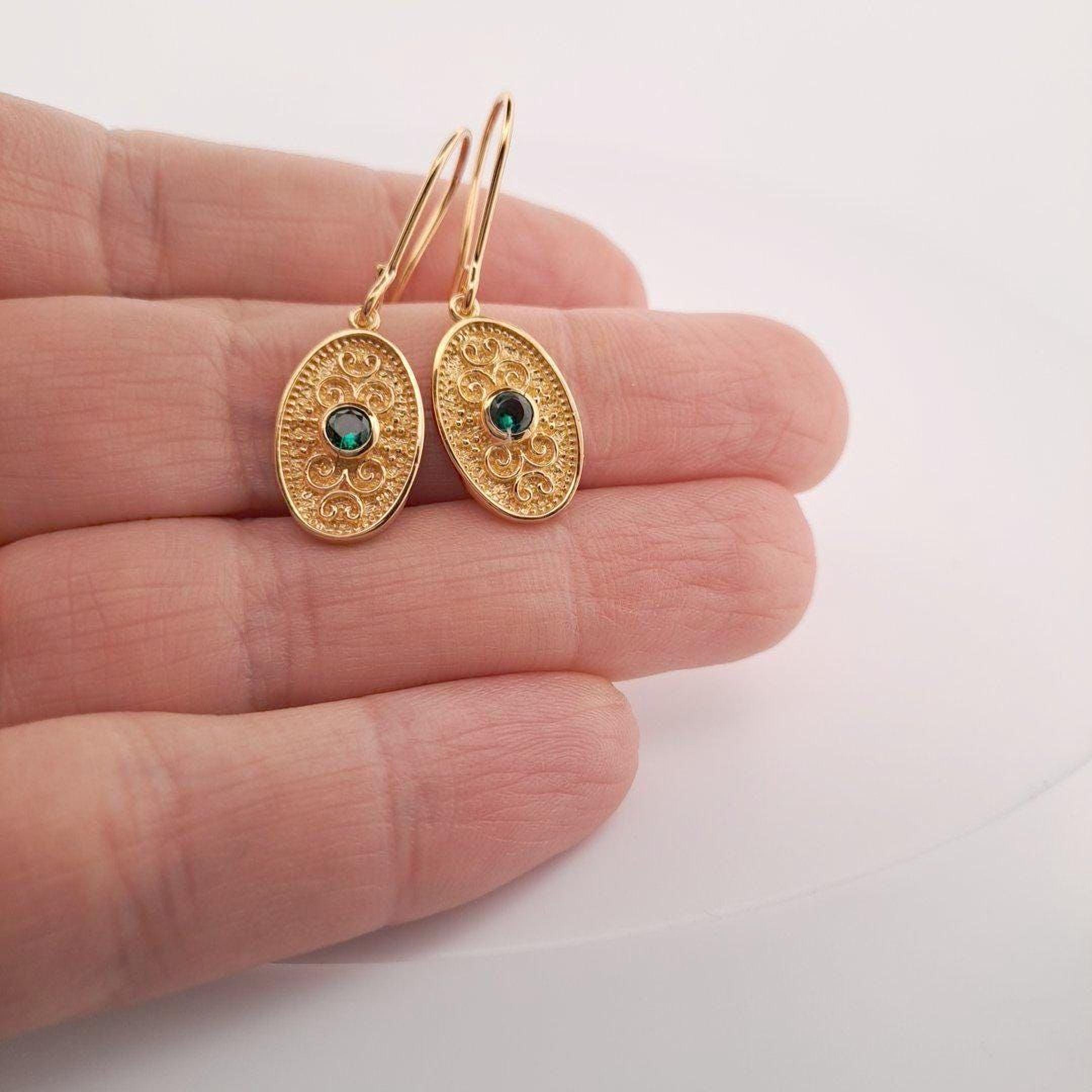 A pair of gold earrings with green gemstones is being held in a hand against a white background.
