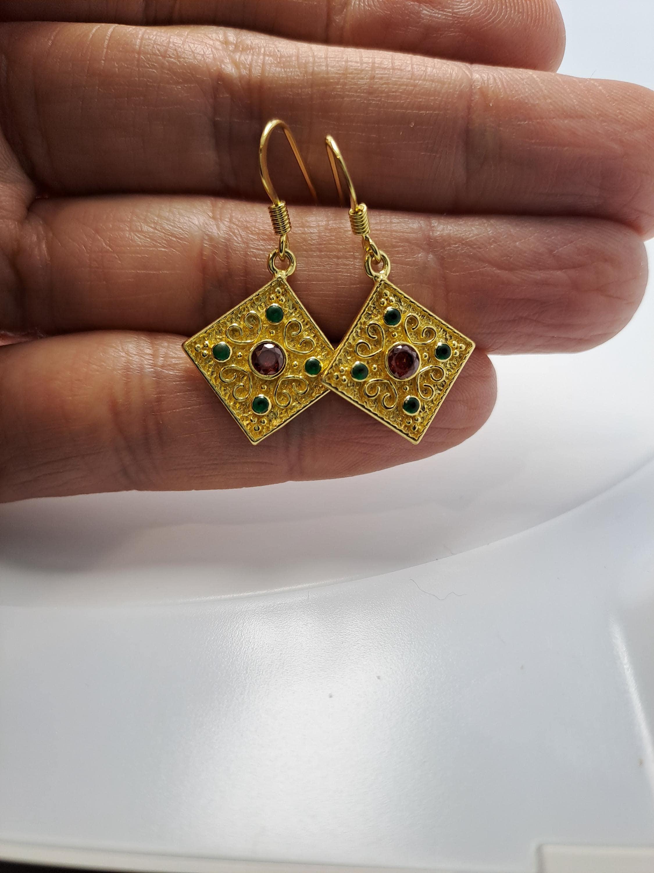 A pair of gold earrings with green and red gemstones, held in a person's hand.