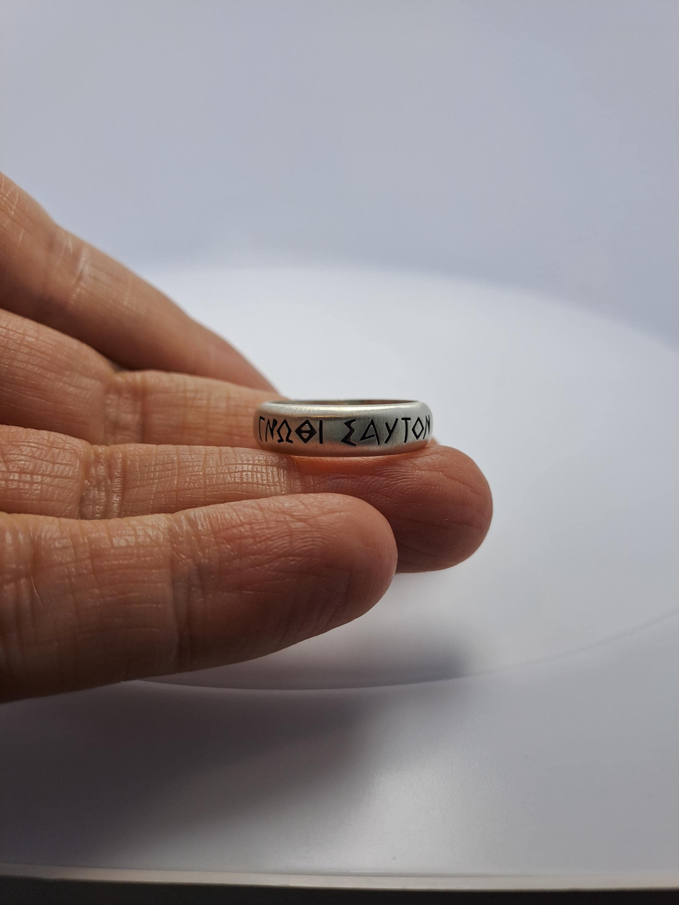 A hand holding a silver ring with Greek text engraved on it.