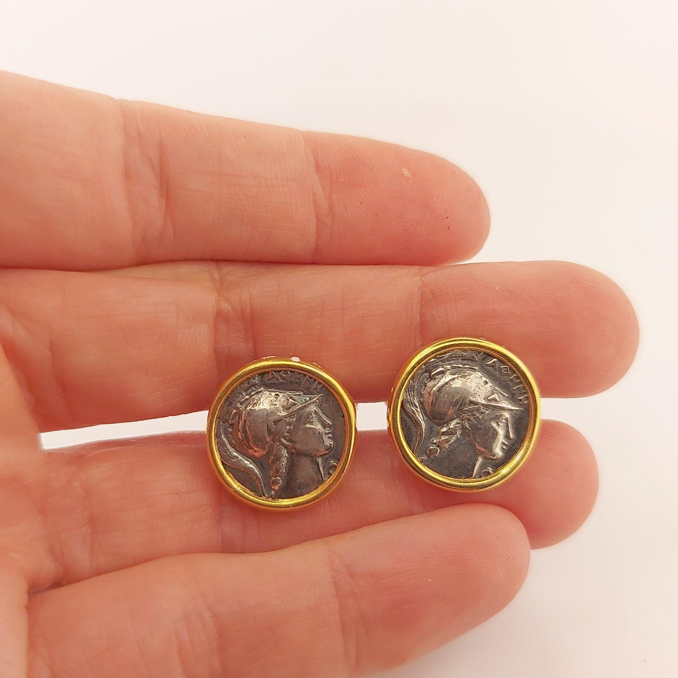 A hand holding two round earrings with a silver and gold design, featuring a profile of a person's face.
