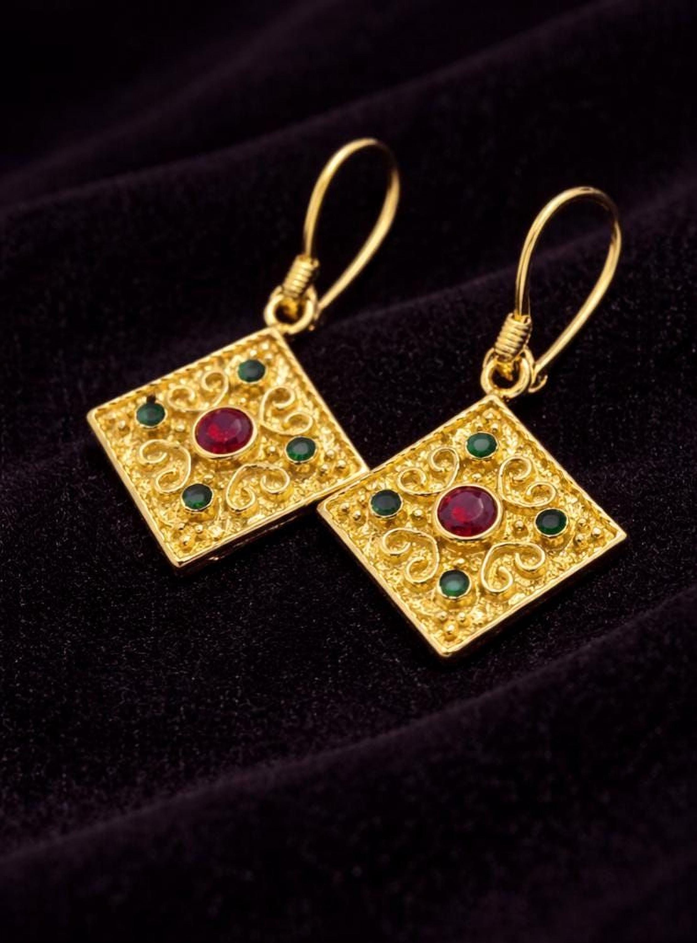 A pair of ornate gold earrings with a red gemstone in the center, set against a dark background.