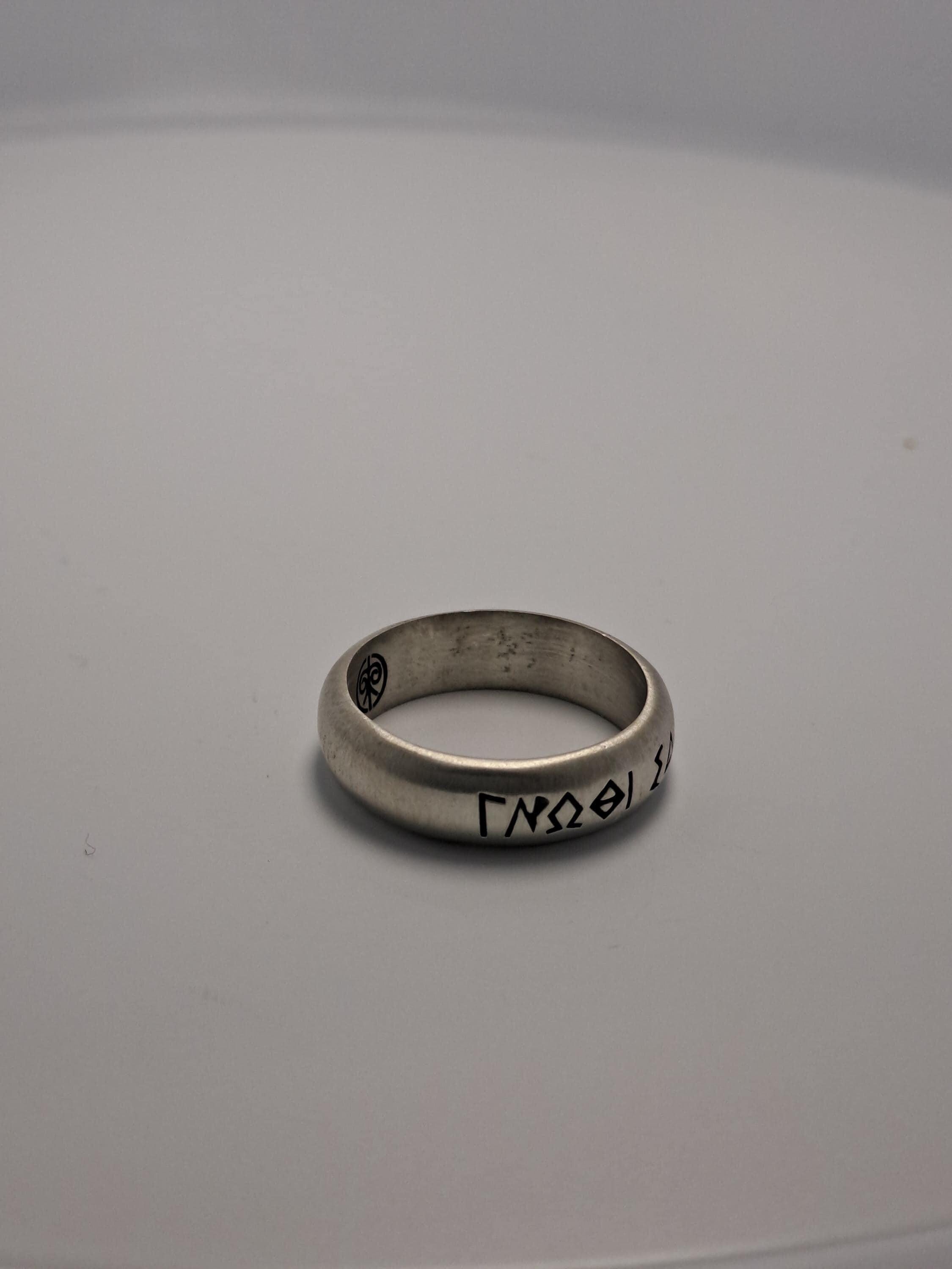 A silver ring with a symbol or text engraved on it, placed on a plain white surface.